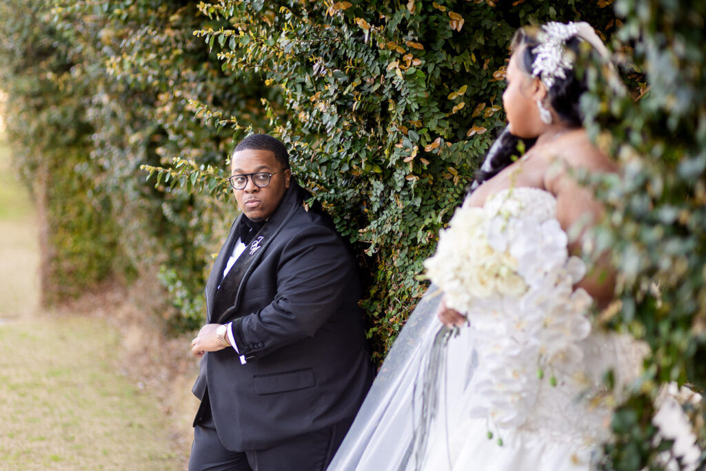 Wedding couple poses against ivy-covered wall outdoors, with person in black suit standing beside flowing white dress.