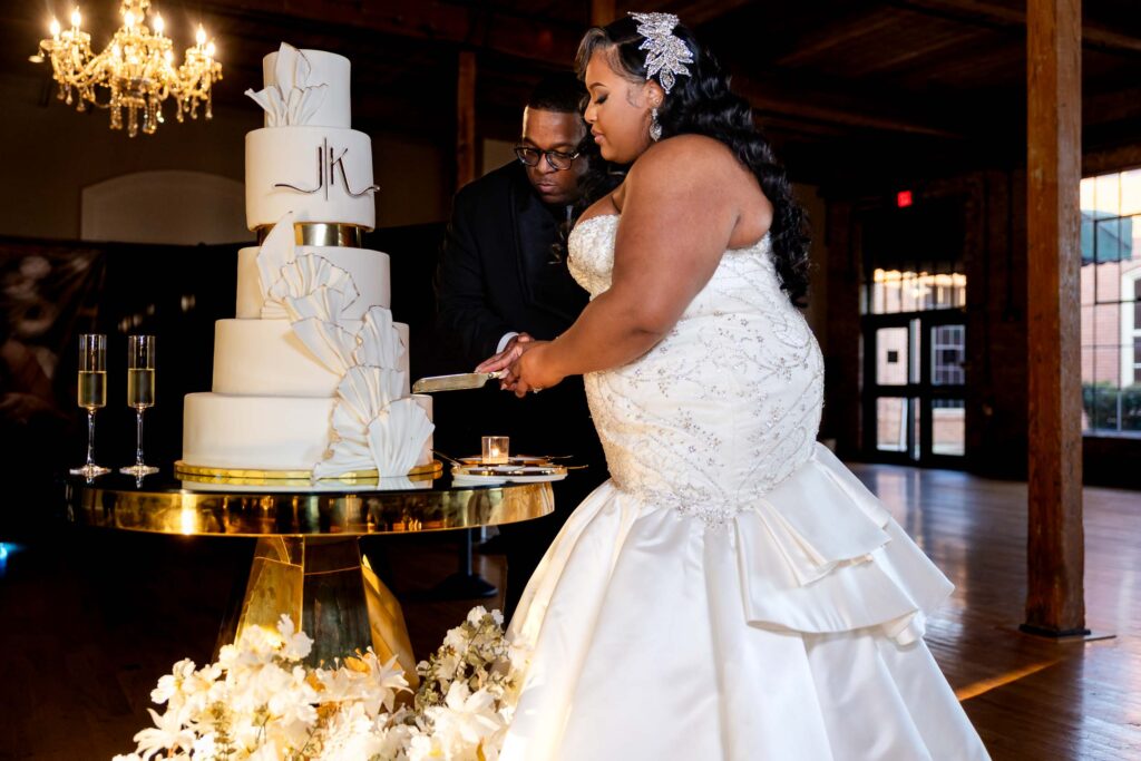 A couple slices into their elegant white wedding cake at their reception, with a crystal chandelier overhead.